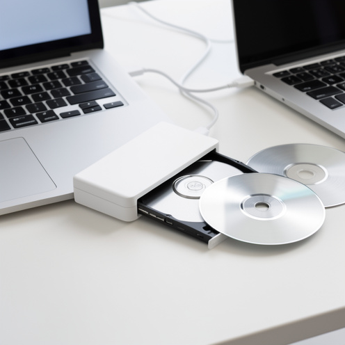Lecteur optique externe blanc connecté à un ordinateur portable par câble USB, avec des disques argentés sur un bureau clair.