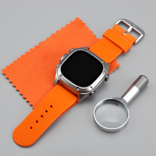 Montre connectée robuste en titane avec bracelet orange, présentée neuve sur un fond gris avec un chiffon et une loupe d’inspection.